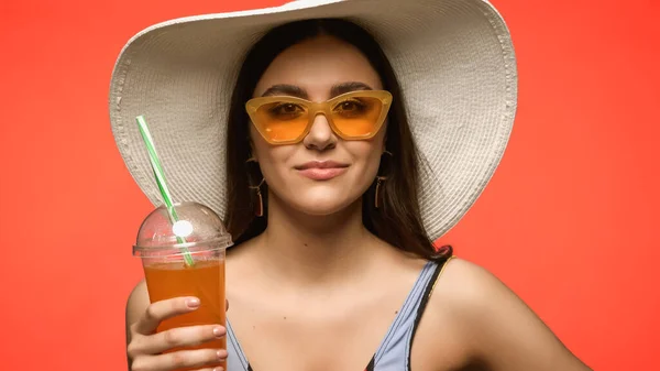 Pretty brunette woman in sun hat holding cocktail in plastic cup isolated on coral
