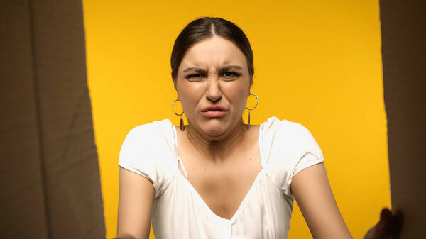 Bottom view of disgusted brunette woman looking at open cardboard box isolated on yellow 