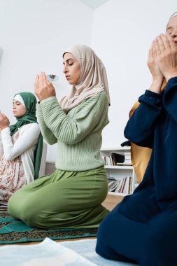 Young muslim woman praying on rug near asian mother and daughter at home  clipart