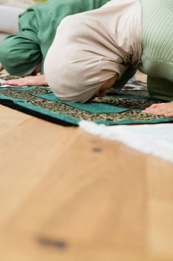 Arabian mother and daughter bending while praying on rugs at home  clipart