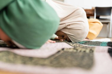 Side view of muslim woman in hijab bending on rug near blurred daughter while praying at home  clipart
