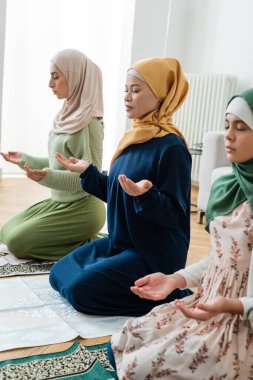 Asian woman in traditional muslim clothes praying near arabian daughter and granddaughter at home  clipart