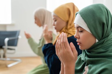 Side view of girl in hijab holding hands near face while praying near multiethnic family  clipart