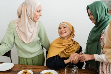 Smiling arabian woman looking at daughter and asian mom near food at home  clipart