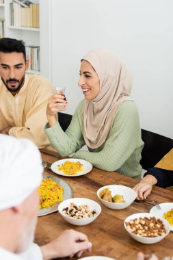 Müslüman ailesiyle yemek yerken tesettürlü neşeli Arap kadın elinde bir bardak çay tutuyor.