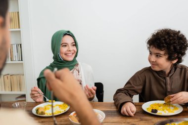 smiling muslim girl pointing with finger during dinner with dad and arabian brother clipart