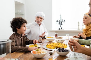 Orta yaşlı Müslüman bir adam, ırklar arası aile yemeği sırasında Arap torununu işaret ediyor.