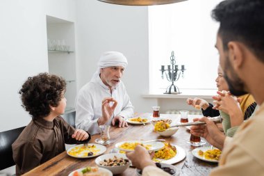 Orta yaşlı Müslüman adam yemek sırasında etnik çeşitliliğe sahip aileyle konuşurken el kol hareketi yapıyor.