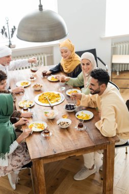 Olgun bir adamın çok kültürlü Müslüman bir ailenin yanına çay dökerek evde yemek yediği yüksek açılı bir manzara.
