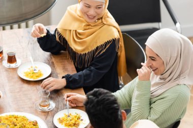 Heyecanlı Müslüman kadının etnik çeşitlilikte bir aileyle yemek yerken gülüşünün yüksek açılı görüntüsü