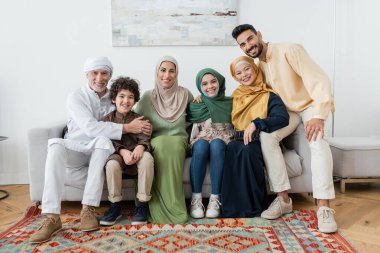 Mutlu çok ırklı Müslüman aile evdeki kanepede kameraya bakıyor.