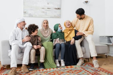 Neşeli, çok kültürlü Müslüman aile evdeki kanepede gülüyor.