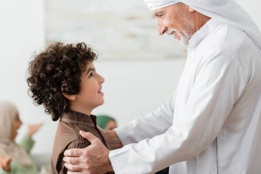 Memlekette Arap torununun omuzlarına sarılan mutlu Müslüman adamın yan görüntüsü.