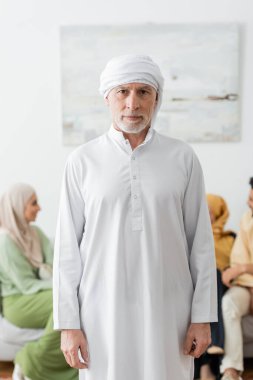 Geleneksel Müslüman kıyafetleri içinde olgun bir adam bulanık ailenin yakınındaki kameraya bakıyor.