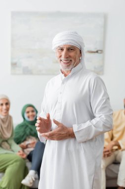Beyaz türbanlı mutlu Müslüman adam bulanık ailenin yakınındaki kameraya bakıyor.