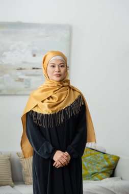 Orta yaşlı tesettürlü Asyalı kadın evde kameraya bakıyor.