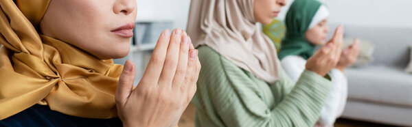 Arabian woman praying near blurred family at home, banner 