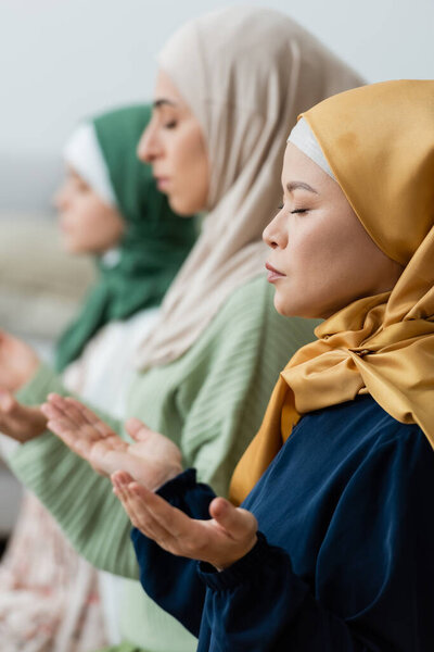 Side view of asian woman in hijab praying near blurred muslim daughter and granddaughter at home 