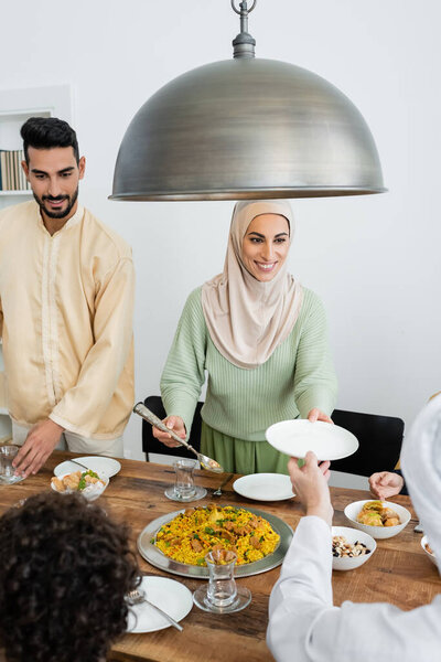 Smiling muslim woman serving pilaf near family and food at home 