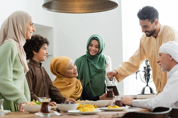 Homem Árabe Derramando Chá Perto Comida Sorrindo Família Interracial Casa — Fotografia de Stock