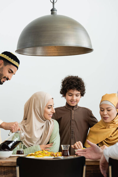 smiling arabian man in skullcap pouring tea near multiethnic family