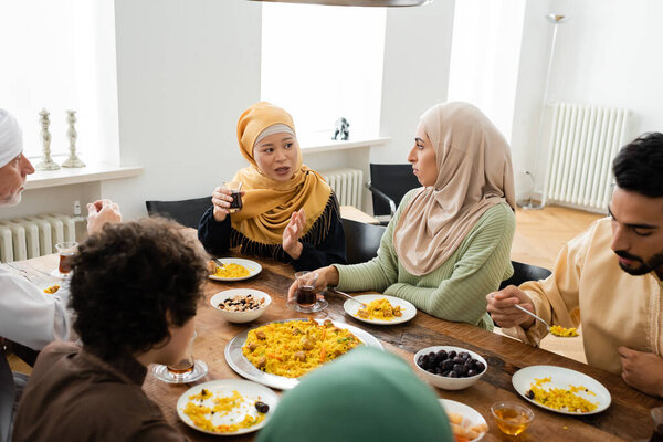 middle aged asian woman pointing with hand while talking to multiethnic muslim family during dinner