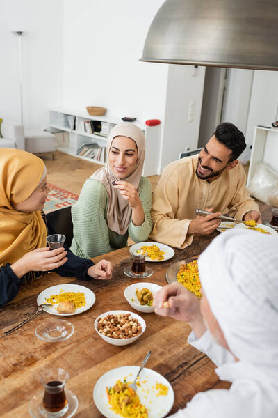 mature asian woman holding glass of tea while talking to muslim family during dinner