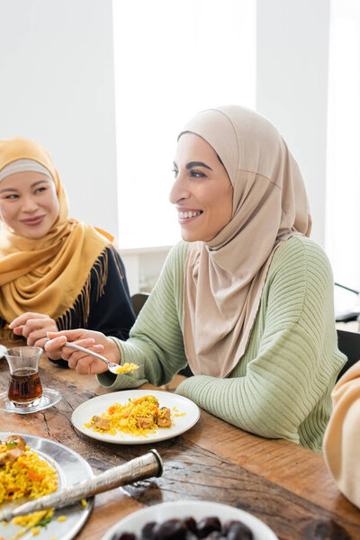 happy muslim woman eating pilaf near asian mother during family dinner