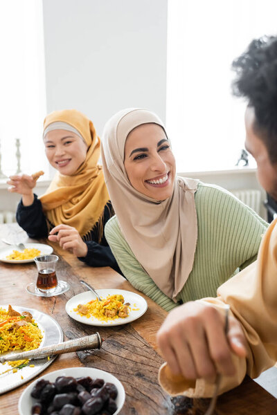 happy muslim woman in hijab smiling near husband and asian woman during dinner