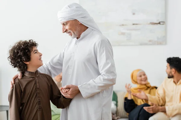 Dojrzały Muzułmanin Trzyma Rękę Arabskiego Wnuka Pobliżu Rozmytej Międzyrasowej Rodziny — Zdjęcie stockowe