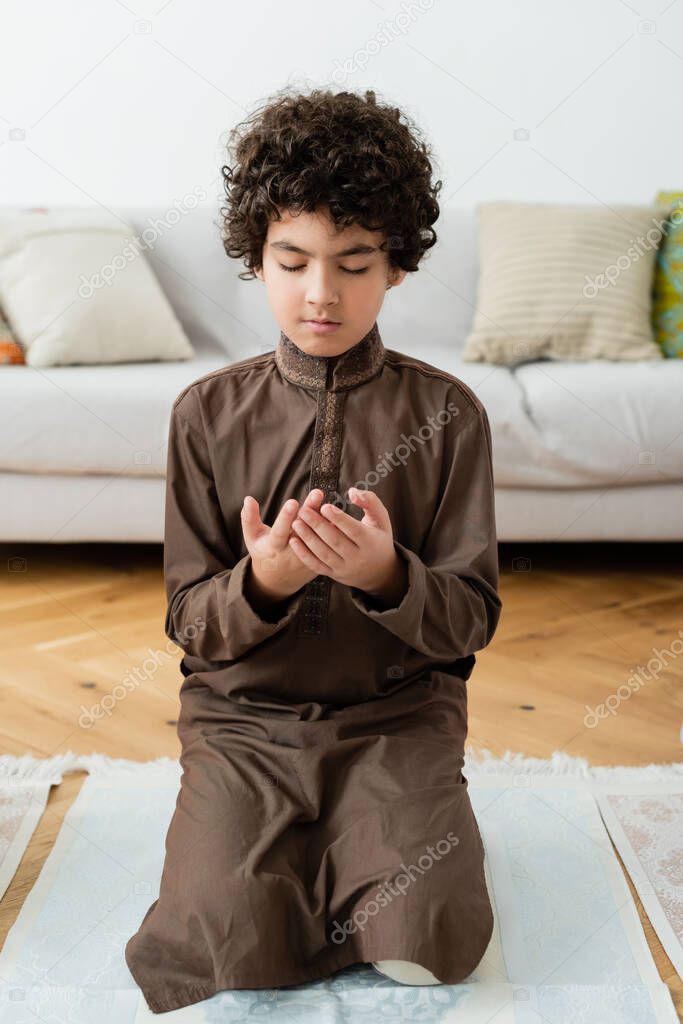 Curly arabian child praying on traditional rug at home 