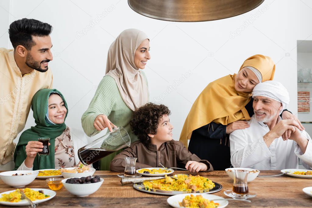 cheerful arabian woman pouring tea during dinner with multiethnic muslim family