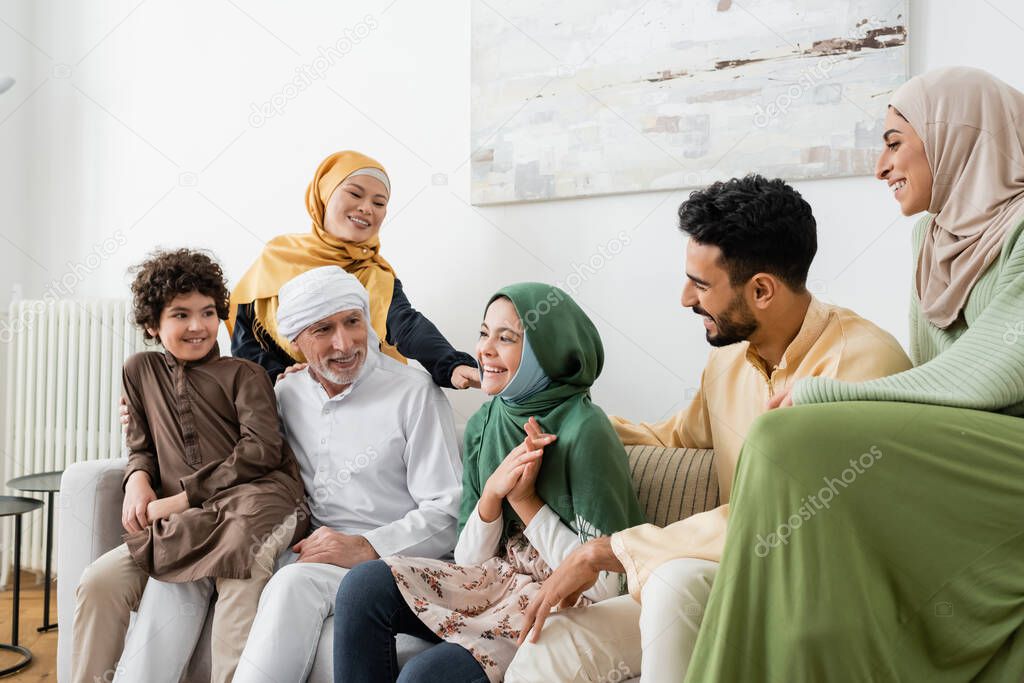happy multicultural muslim family looking at smiling girl while sitting on couch at home