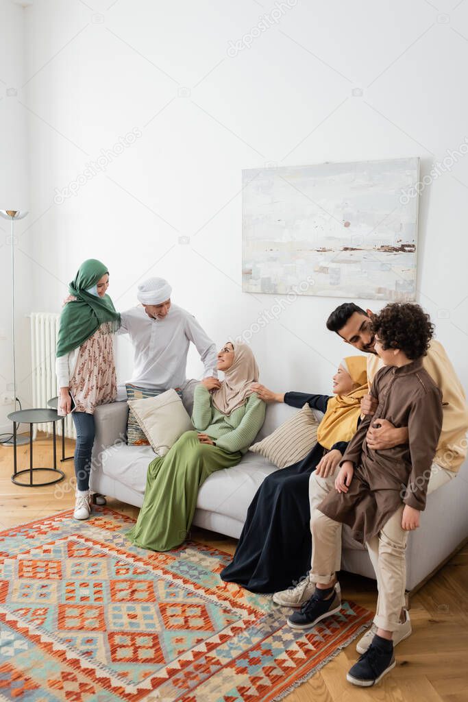 young muslim man hugging arabian son near cheerful multicultural family in living room
