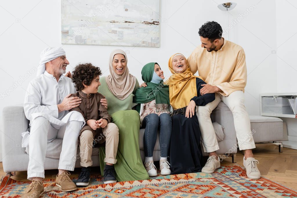 joyful multicultural muslim family laughing on sofa at home