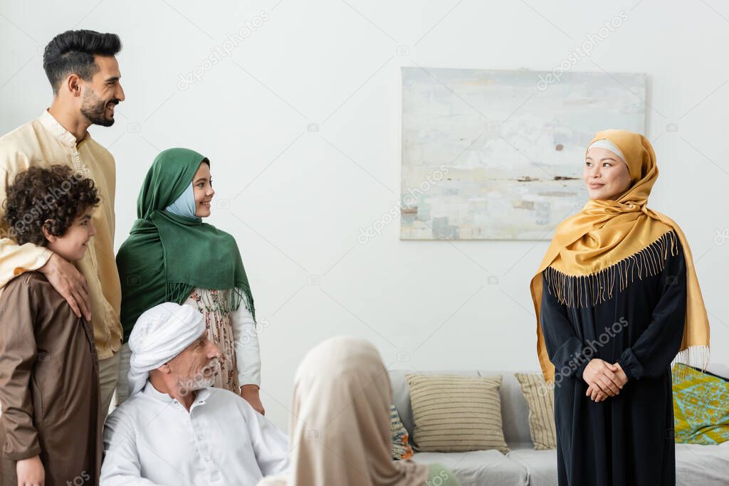 smiling asian woman in hijab looking at multiethnic muslim family at home