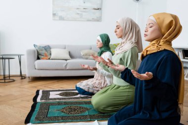 multiethnic muslim women in traditional clothes praying on rugs in living room clipart