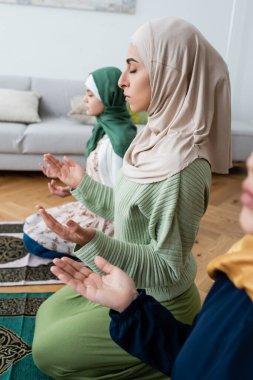 young muslim woman with closed eyes praying near mother and daughter at home clipart