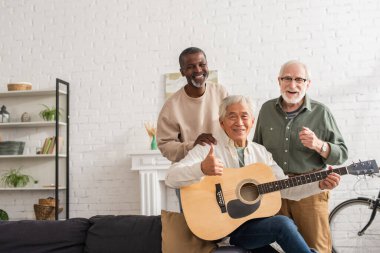 Gülümseyen Asyalı adam akustik gitar tutuyor ve evdeki ırklar arası arkadaşları gösteriyor. 