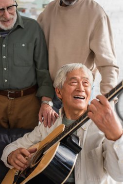 Yaşlı Asyalı adam evdeki ırklar arası arkadaşlarının yanında akustik gitar çalıyor. 