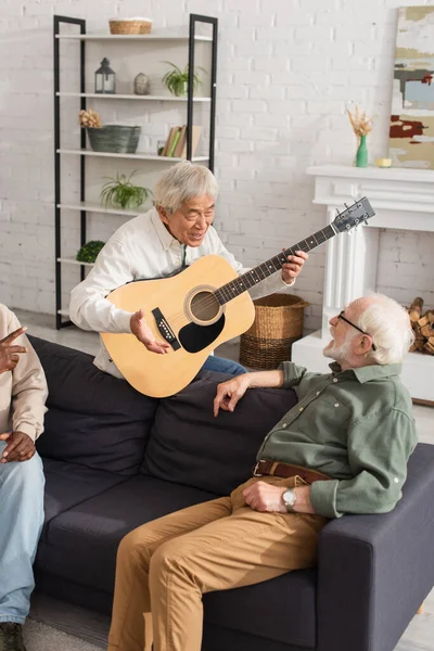 Senior asian man holding acoustic guitar while talking to friend on couch at home