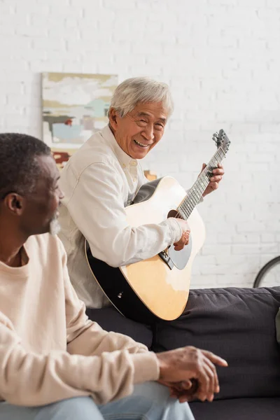 Sonriente Asiático Hombre Jugando Guitarra Acústica Cerca Borrosa Africano Americano — Foto de Stock