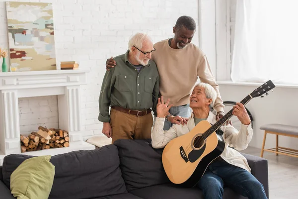 Smiling Multiethnic Men Hugging Asian Friend Acoustic Guitar Home — Stock Photo, Image