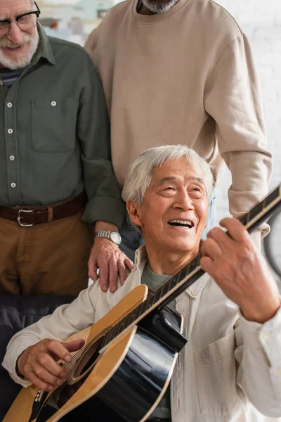 Elderly asian man playing acoustic guitar near interracial friends at home