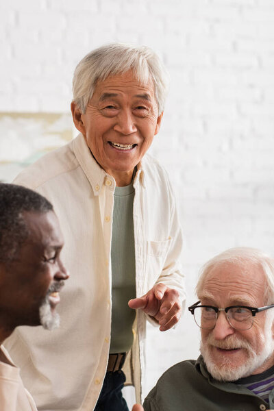 Happy asian man looking at camera near multiethnic friends at home 