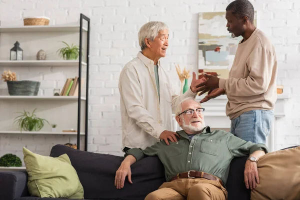 Homens Multiétnicos Conversando Perto Amigo Sênior Sofá Casa — Fotografia de Stock