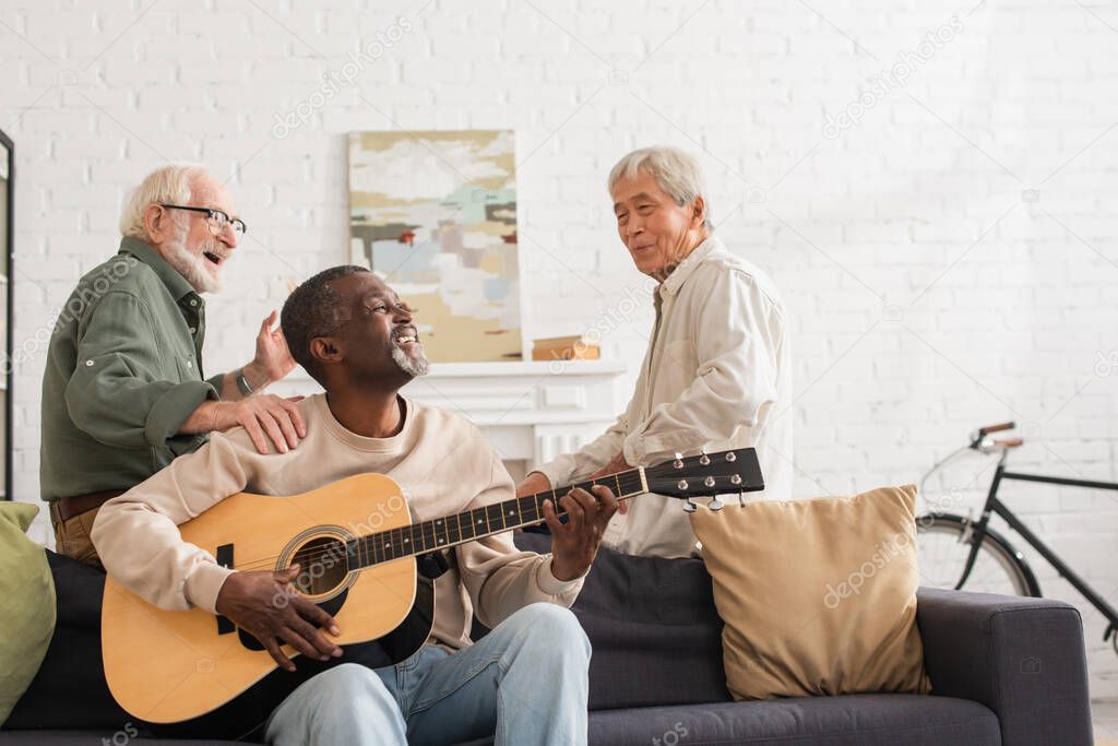 African american man playing acoustic guitar near multiethnic friends in living room 
