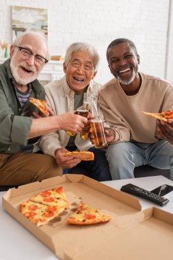 Evdeki uzaktan kumandanın yanında pizza ve bira tutan mutlu ırklar arası yaşlılar. 