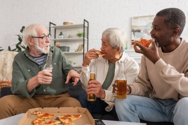 Bira şişesi tutan ve oturma odasında pizza yiyen ırklar arası arkadaşlarla konuşan yaşlı bir adam. 