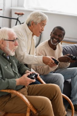 KYIV, UKRAINE - 12 Ekim 2021: Odaklanmış Asyalı adam evde yaşlı arkadaşlarıyla video oyunu oynuyor 